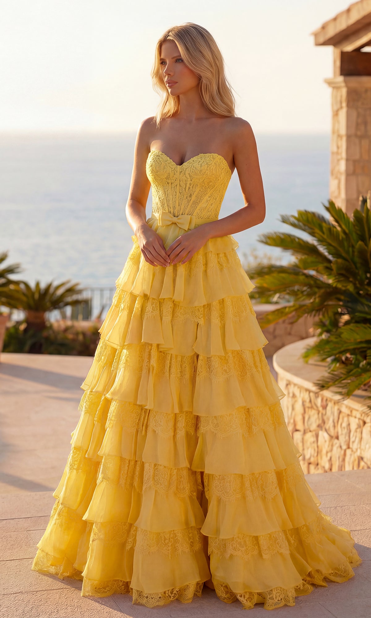 A woman stands outdoors in a yellow Chiffon and Lace Prom Gown: PromGirl Exclusive 250238. Elegant stone architecture, lush plants, and an ocean view provide a stunning backdrop for this strapless tiered prom dress.
